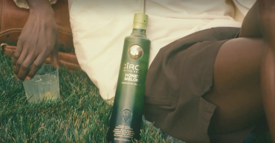 A bottle of Cîroc Honey Melon resting on the grass beside a seated figure and a glass