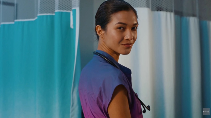 A nurse in ombre FIGS scrubs looks back over her shoulder by hospital curtains