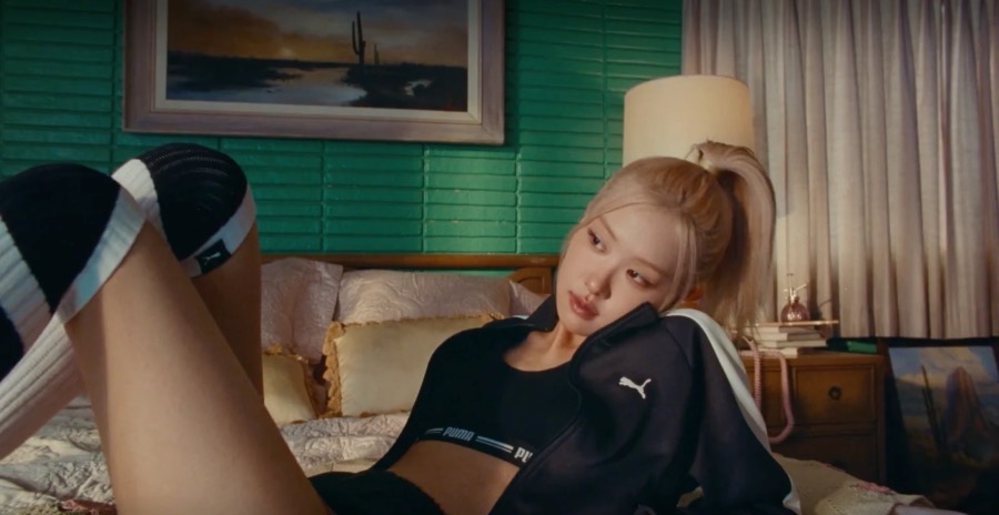 A woman in a black Puma track jacket lounges on a bed in a retro green motel room