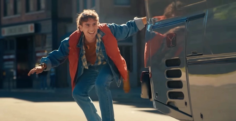 A skateboarder in a red vest and denim grabs onto a moving silver car